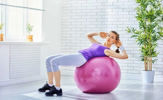 picture of a woman on an exercise ball on a wooden floor beside a window and flower on the other side 